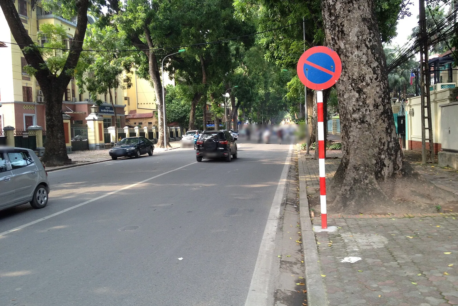 Biển P.131a tròn viền đỏ nền xanh một vạch chéo cấm đỗ xe, cắm trên cột sơn đỏ trắng bên vỉa hè phố Chu Văn An, Hà Nội, ô tô và xe máy chạy phía trước